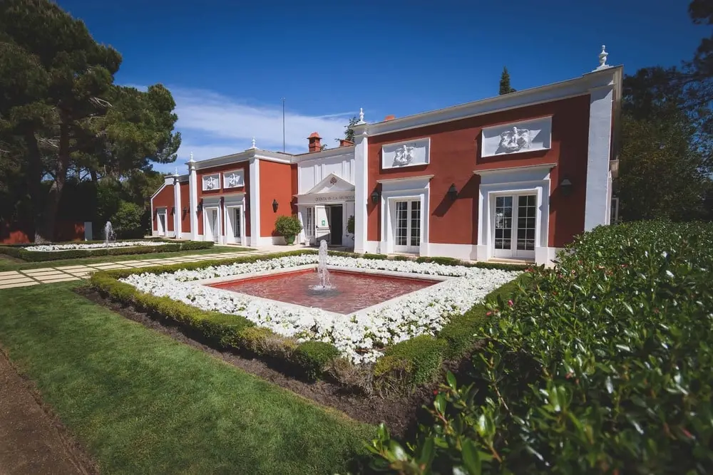 La casa roja de columnas blancas y el parterre de la fuente, bajo un cielo azul de verano