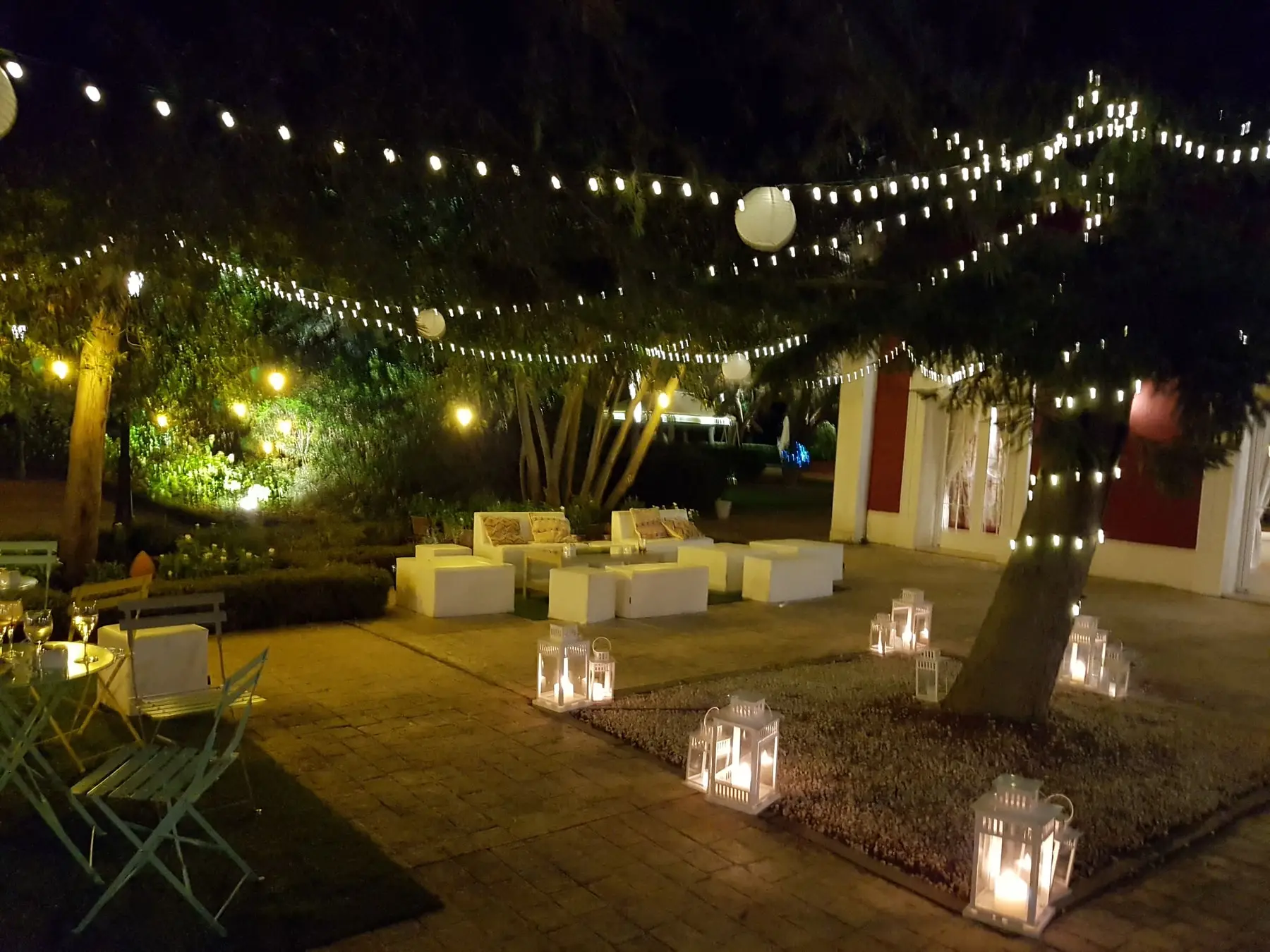 El jardín de noche: guirnaldas de bombillas entre los árboles, sofás blancos y farolillos