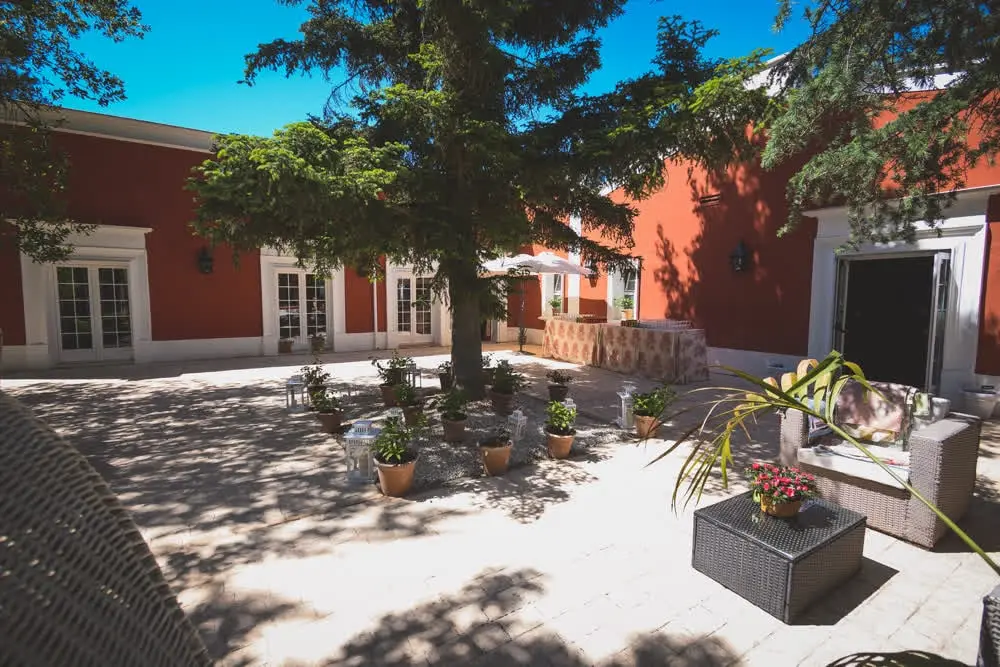 Patio de losas a la sombra de un árbol, entre muros rojos y ventanas blancas